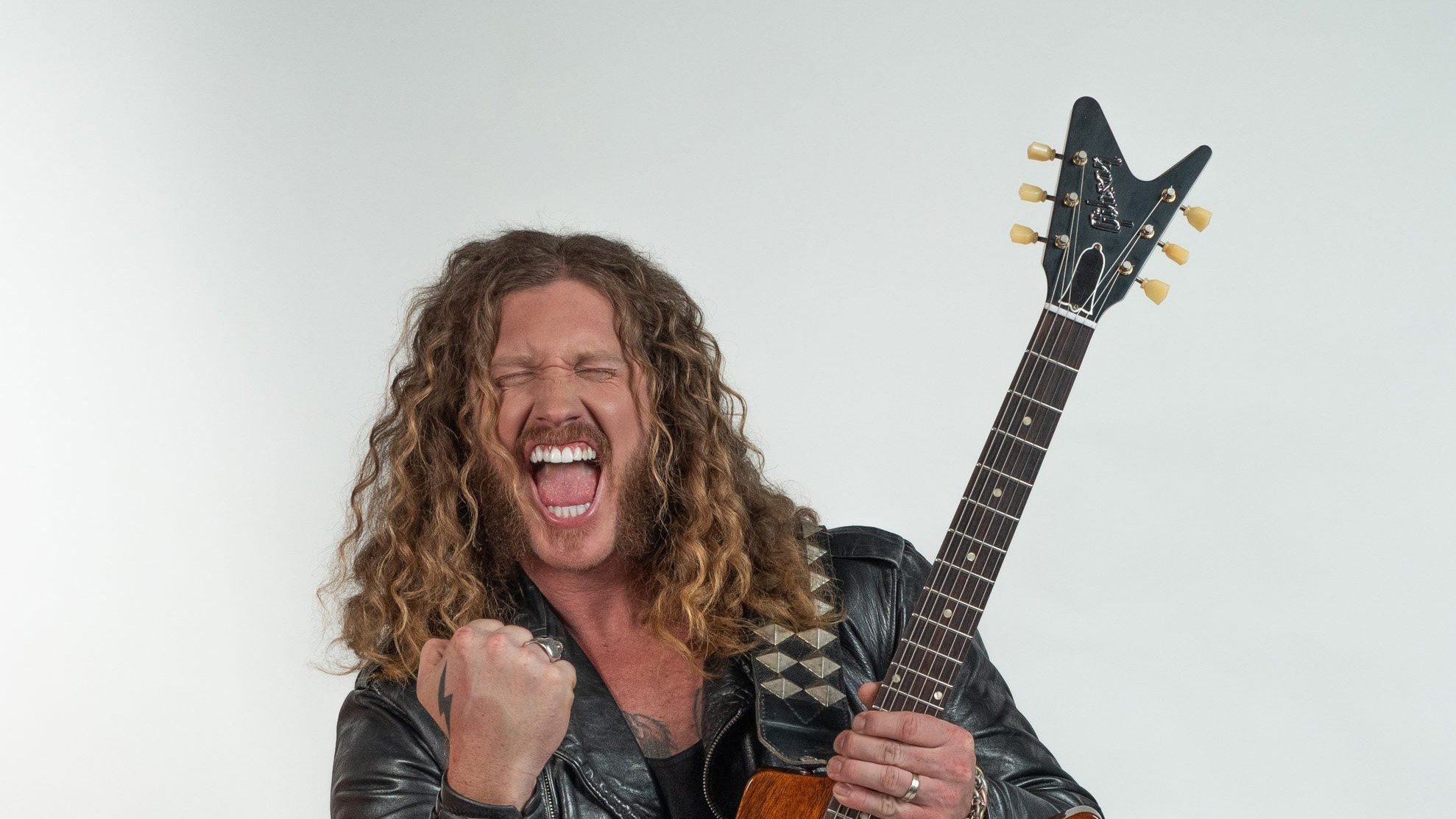 Jared James Nicols mit langer Lockenmähne und E-Gitarre vor hellem Hintergrund, gekleidet in schwarze Lederjacke und Jeans. Die Person hält die Faust erhoben und präsentiert die Gitarre in einer dynamischen Pose.