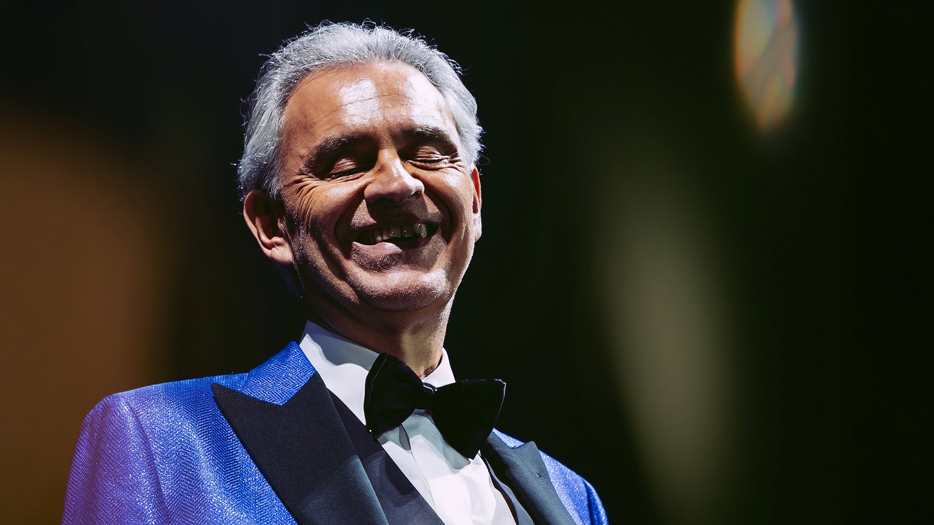 Andrea Bocelli in einem blauen Anzug mit weißem Hemd und schwarzer Fliege lachend vor dunklem Hintergrund mit Lichtreflexen