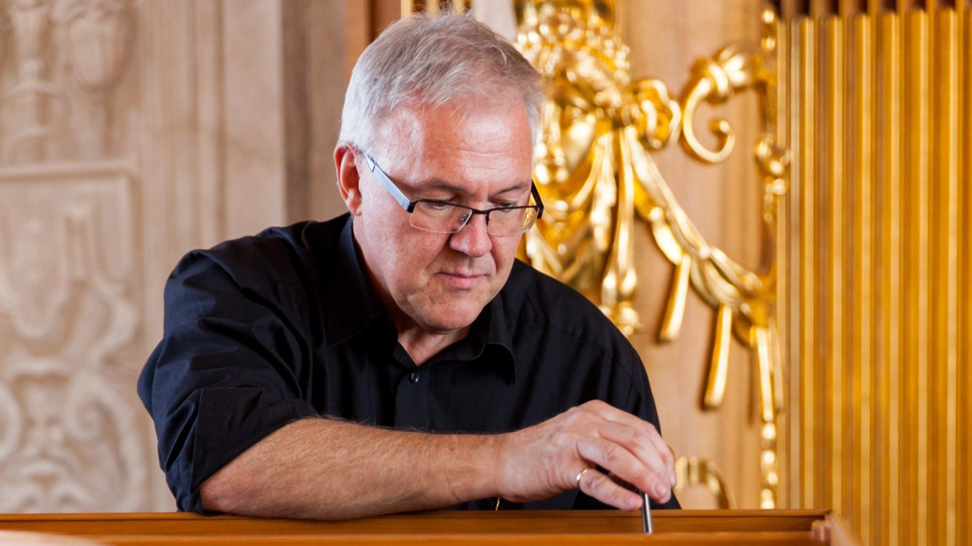 Person mit Brille an einer historischen Orgel in einem barocken Innenraum. Vergoldete Verzierungen und Orgelpfeifen prägen den Hintergrund, während eine Hand an den Bedienelementen des Instruments liegt