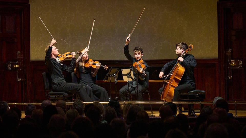 Vier Musiker mit Instrumenten, dunkel gekleidet auf Bühne
