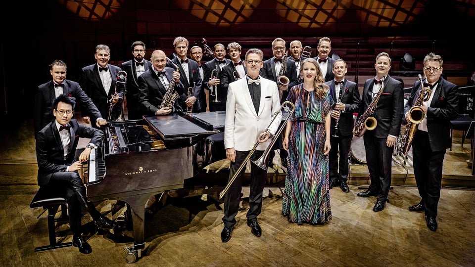 Eine Bigband in formeller Kleidung steht auf einer Konzertbühne; im Vordergrund ein Flügel sowie zwei Musiker*innen mit Posaune und Mikrofon, dahinter weitere Bläser des Ensembles.
