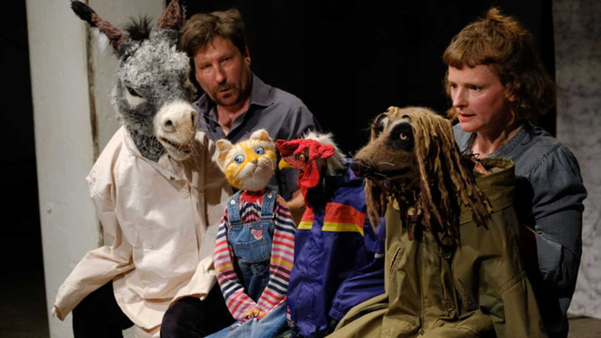 Bühnenfoto aus Die Bremer Stadtmusikanten mit zwei Darstellenden und mehreren Tierfiguren, darunter ein Eselkopf, eine Katzenpuppe, ein Hahn und eine hundeähnliche Figur, auf einer Theaterbühne vor dunklem Hintergrund.