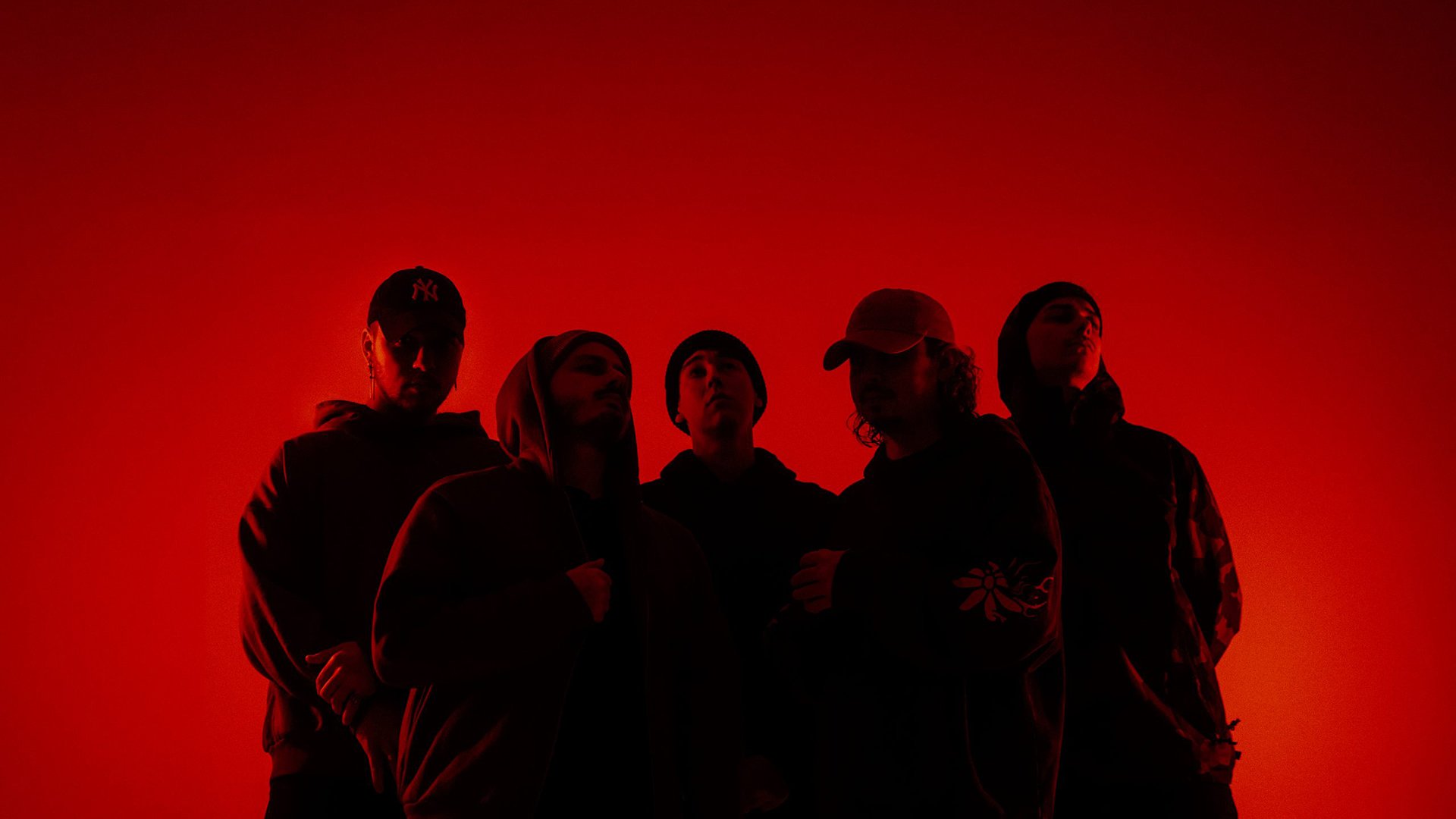 Bandfoto von Accvsed mit fünf Bandmitgliedern in dunkler Kleidung vor leuchtend rotem Hintergrund, darunter Personen mit Kapuzenpullovern, Mützen und Kappen in einer dicht stehenden Gruppenformation.