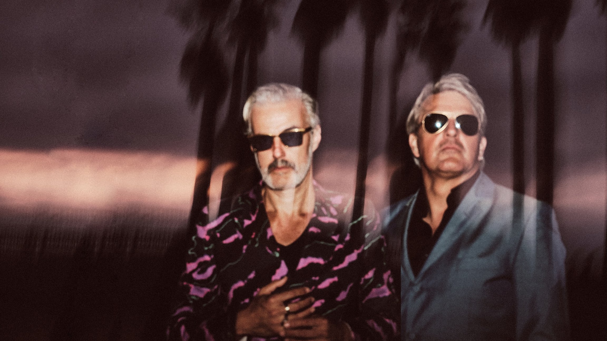 Zwei Personen von Triggerfinger mit Sonnenbrille, einer mit gemustertem Hemd, im Hintergrund verschwommene Palmen im Dunkeln