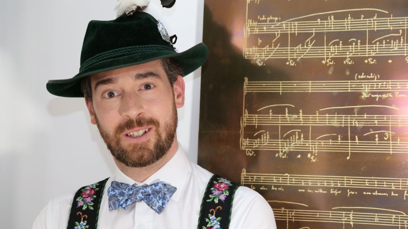 Porträt eines Mannes in bayerischer Tracht mit Hut und Fliege vor einer Tafel mit Notenschrift.
