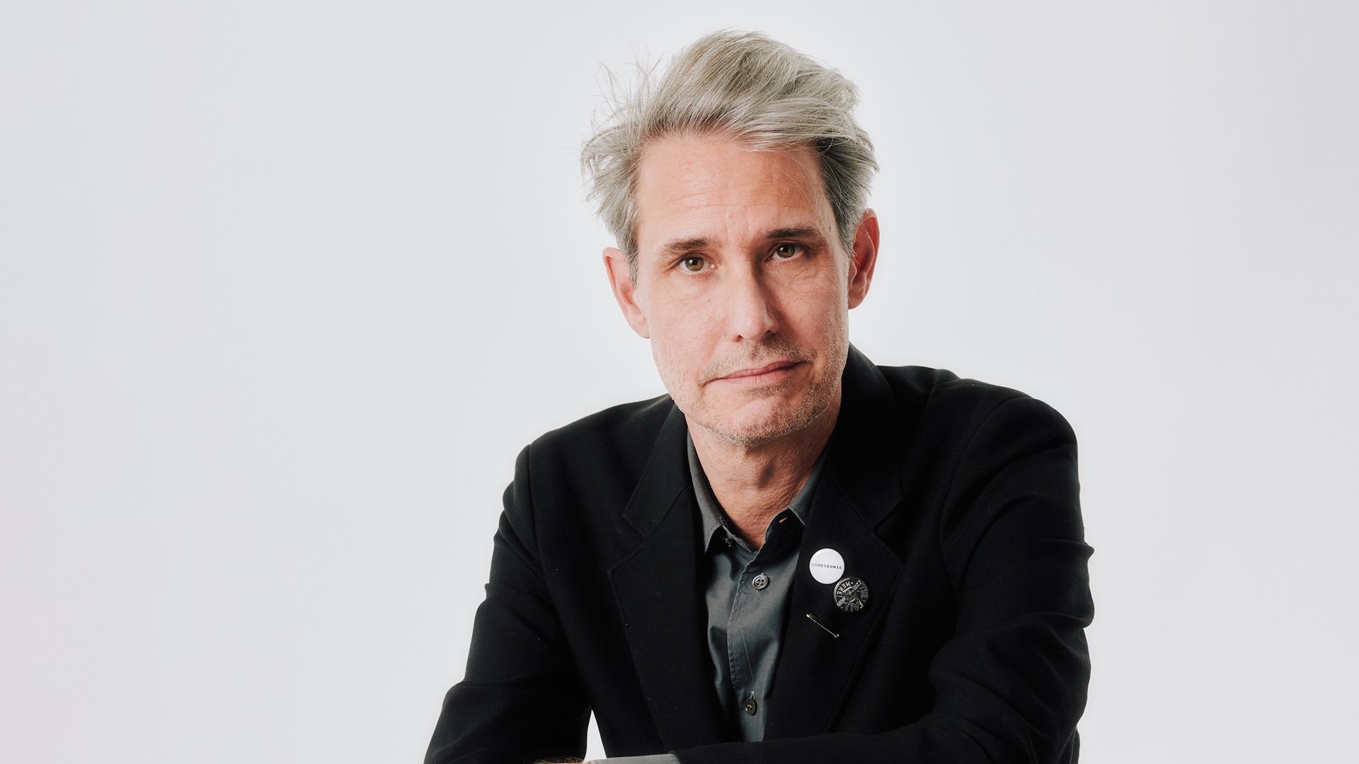Porträt einer Person mit grauem Haar in dunklem Sakko vor hellem Hintergrund. Minimalistische Studioaufnahme mit direktem Blick in die Kamera.