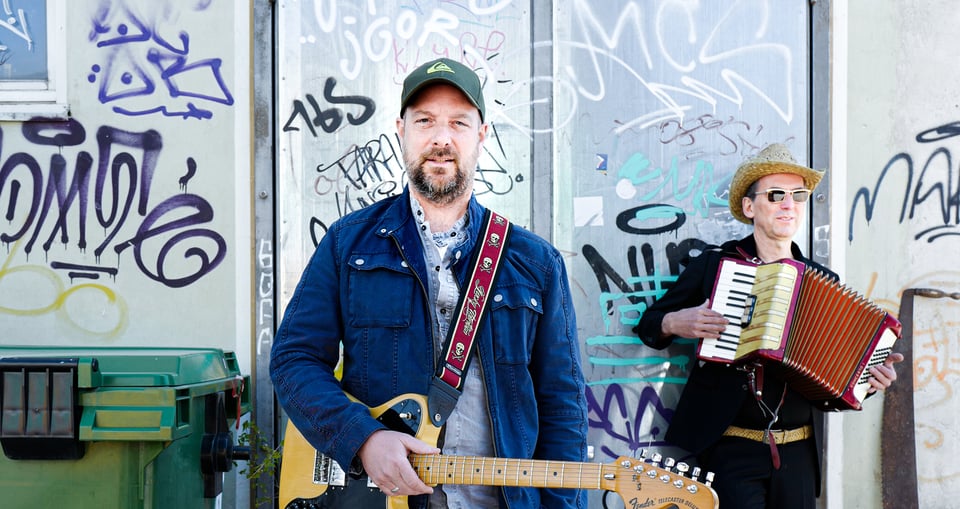 Bild von Künstler mit Gitarre und weiterer Person mit Akkordeon vor Graffiti Wand 