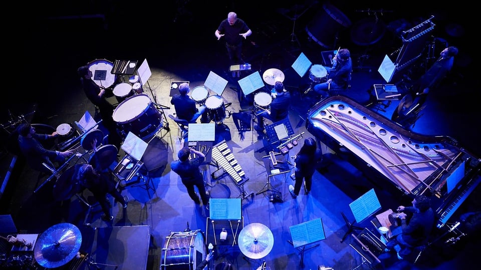 Ein Ensemble von MusikerInnen mit Schlaginstrumenten und Klavier, angeordnet im Kreis auf einer Bühne; eine dirigierende Person steht in der Mitte, Notenpulte sind verteilt 