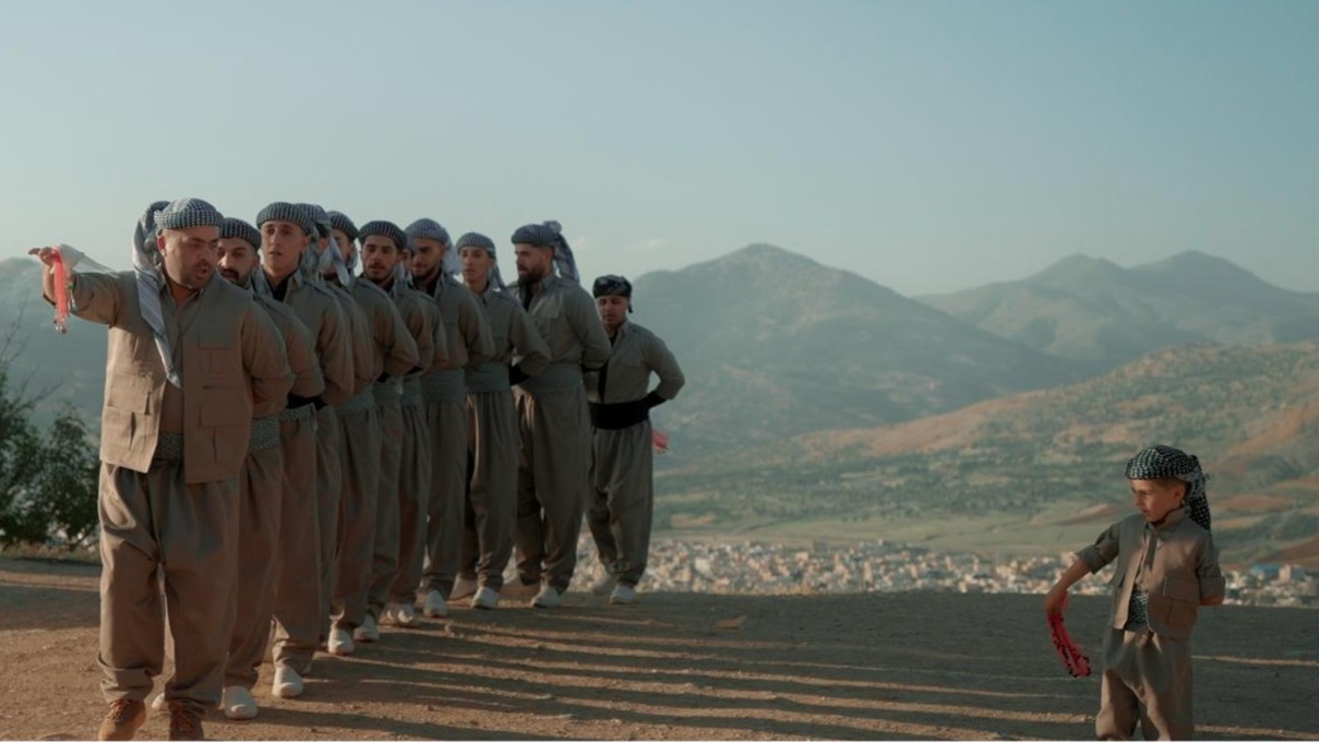 Mehrere Personen stehen in traditioneller Kleidung in einer Reihe und führen einen Reigentanz auf einer Anhöhe vor bergiger Landschaft aus; im Vordergrund bewegt sich ein Kind mit einem roten Tuch.