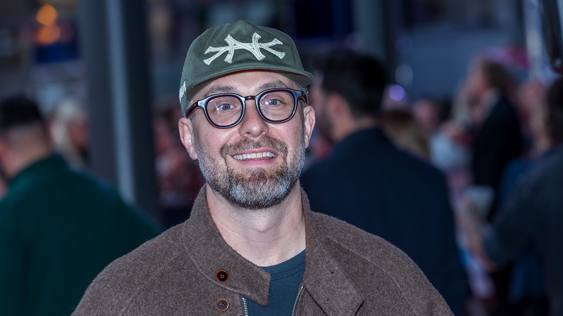 Porträtfoto von Mark Forster mit Brille, grüner Kappe und brauner Jacke vor unscharfem Veranstaltungsumfeld, frontal zur Kamera blickend.