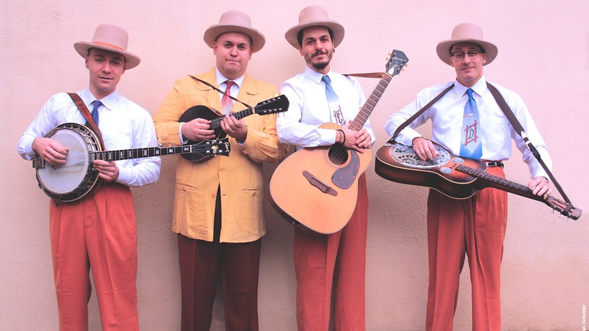Die vier Mitglieder der Truffle Valley Boys mit Gitarren in farbiger Kleidung und rosa Hüten vor rosa Hintergrund
