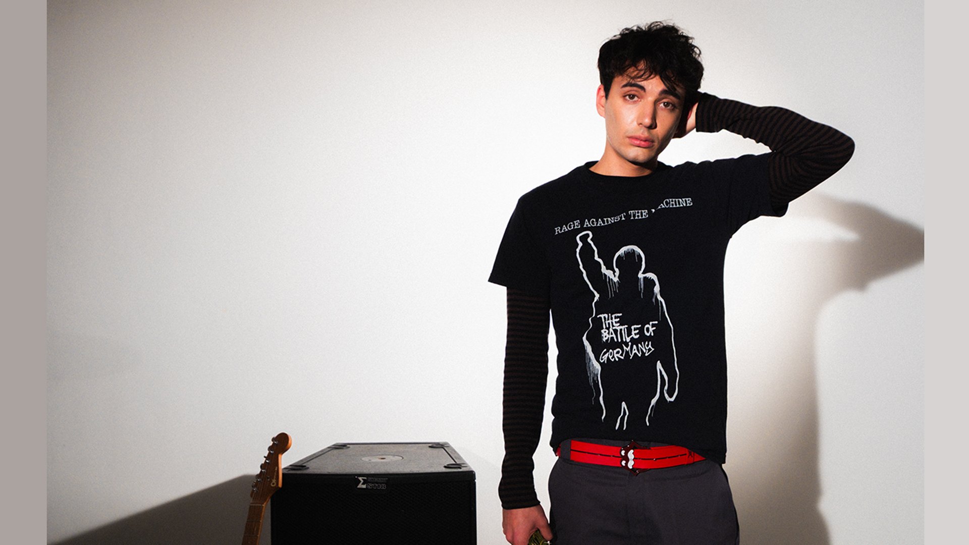 Ein junger Mann mit dunklen Haaren und schwarzem Tshirt mit weißem Aufdruck und rotem Gürtel neben einem Verstärker und einer Gitarre stehend