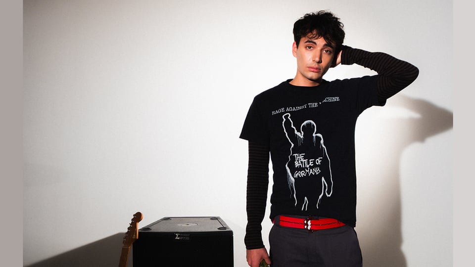 Ein junger Mann mit dunklen Haaren und schwarzem Tshirt mit weißem Aufdruck und rotem Gürtel neben einem Verstärker und einer Gitarre stehend