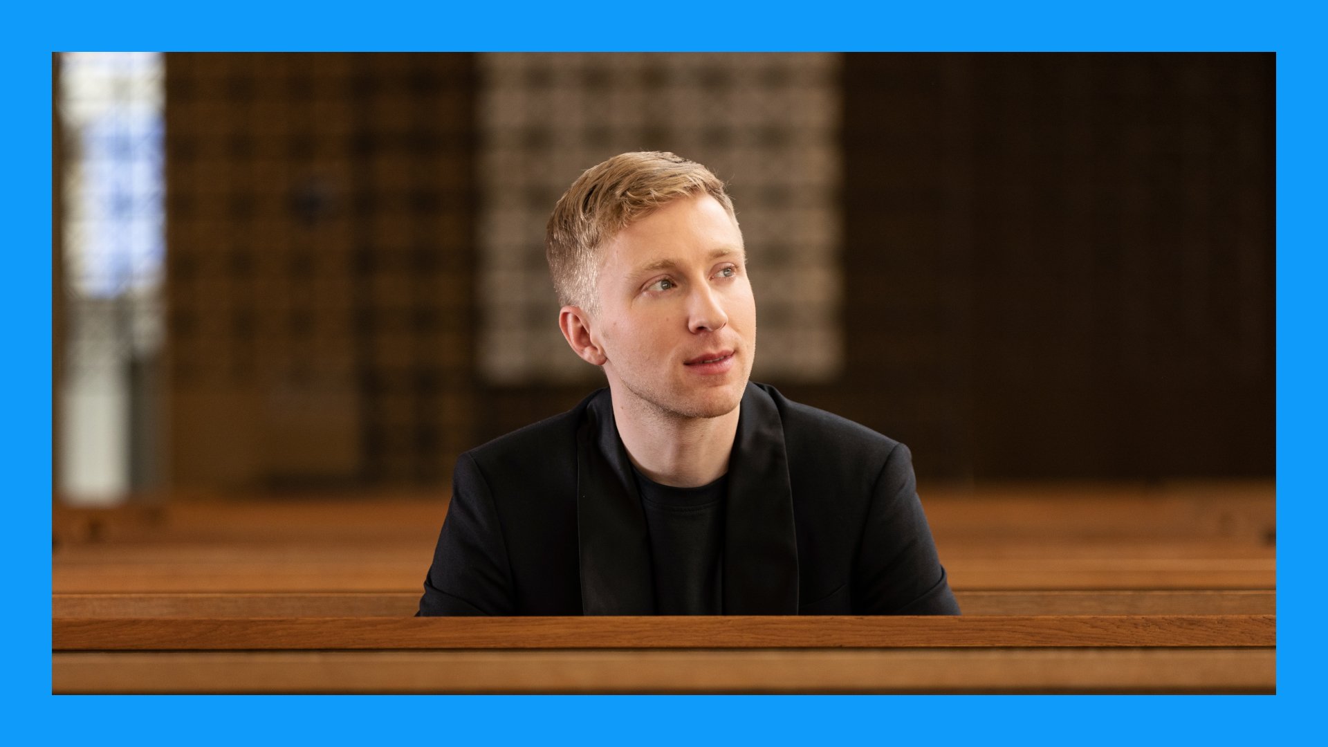 Patrick Hahn an einem Holzpult, zur Seite Blickend, das Bild hat einen blauen Rahmen