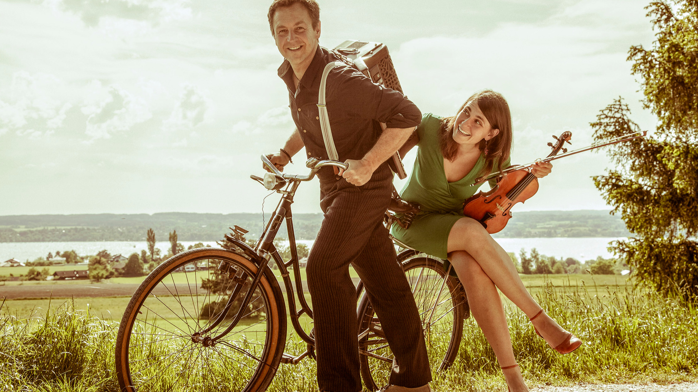 Ein Mann mit Fahrrad und eine Dame mit Instrument auf dem Gepäckträger sitzend 