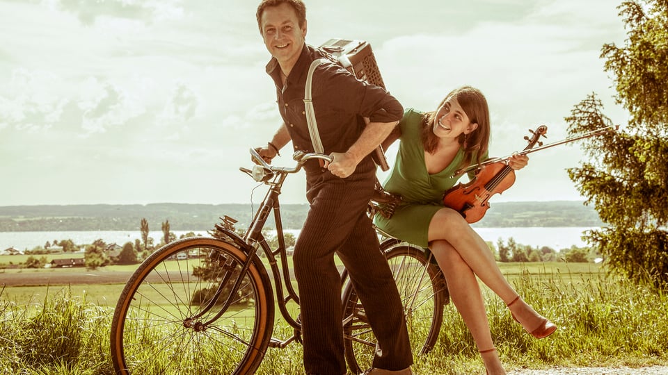 Ein Mann mit Fahrrad und eine Dame mit Instrument auf dem Gepäckträger sitzend 
