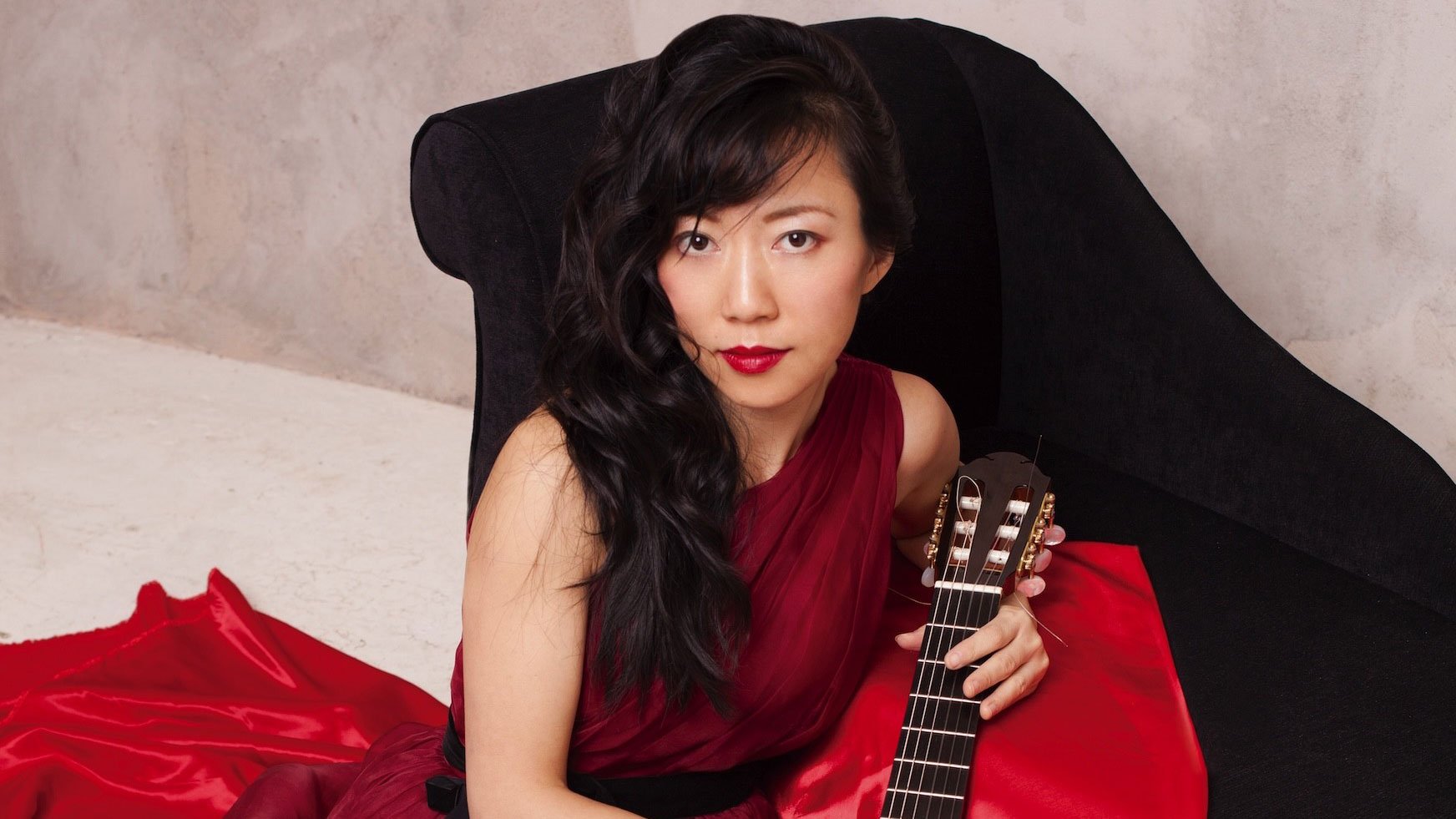 Person in dunkelrotem Kleid sitzt in einem schwarzen Sessel und hält den Hals einer klassischen Gitarre. Dunkles gewelltes Haar, roter Lippenstift und heller Hintergrund prägen die Porträtaufnahme.