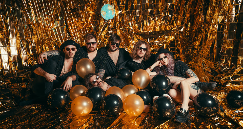 Foto der Band, sitzend auf dem Boden mit orangefarbenen und schwarzen Luftballons, umgeben von glänzend orangener Folie an Wand und Boden.
