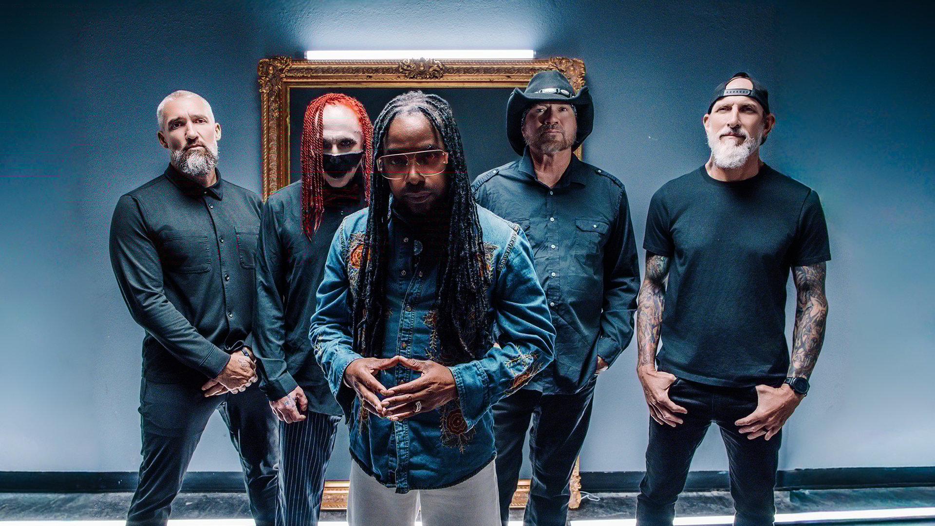 Die fünf Mitglieder der Band Sevendust in dunkler Kleidung stehen frontal in einem Raum vor dunkelblau-grauer Wand, mittig eine Person mit langen Haaren im Vordergrund, im Hintergrund ein beleuchteter, antiker Bilderrahmen