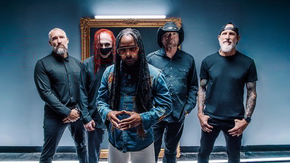 Die fünf Mitglieder der Band Sevendust in dunkler Kleidung stehen frontal in einem Raum vor dunkelblau-grauer Wand, mittig eine Person mit langen Haaren im Vordergrund, im Hintergrund ein beleuchteter, antiker Bilderrahmen