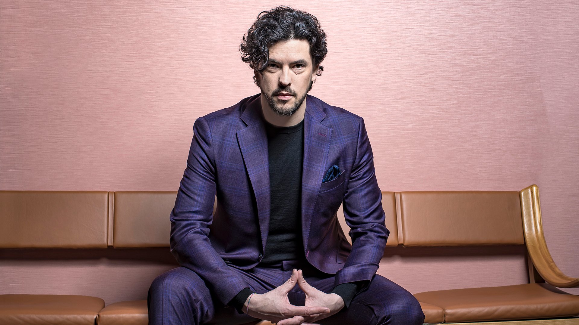 Porträtmotiv zur Veranstaltung mit Andrè Schuen: Sitzende Person in einem violettfarbenen Anzug auf einer gepolsterten Bank vor rosafarbenem Hintergrund. Die Hände sind vor dem Körper zusammengeführt, der Blick ist frontal ausgerichtet. Ruhige Studio‑Beleuchtung und klare Linien; elegantes Promofoto für einen klassischen Liederabend (Gesang & Klavier).