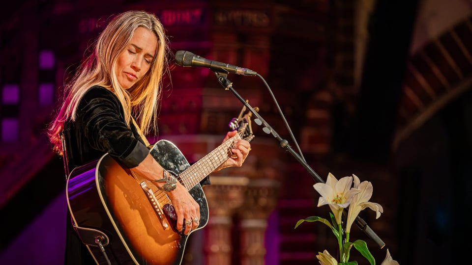 Heather Nova mit Konzertgitarre auf der Bühne  in schwarzer kleidung und offenem Haar, in Vordergrund eine Blume