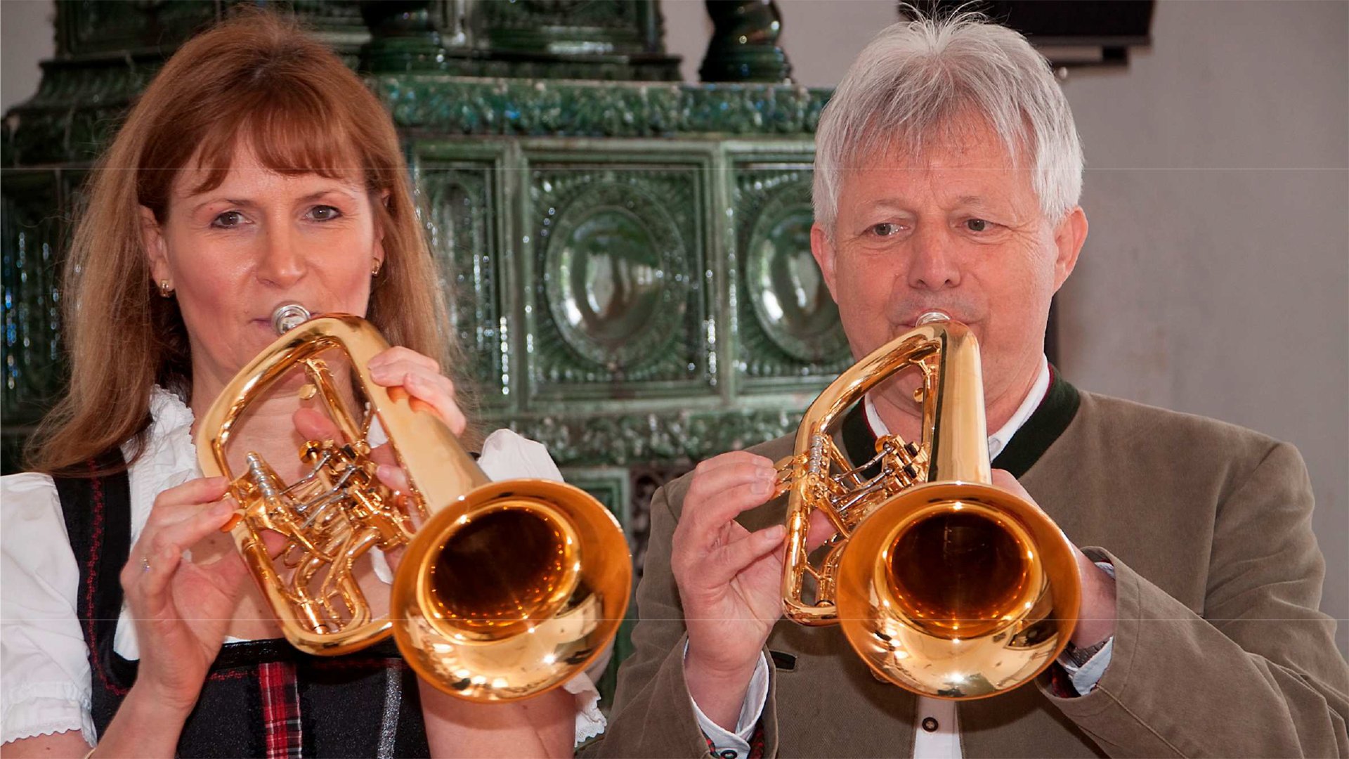 Auftritt von Ingeborg Lutz und Franz L. Schachtner: Zwei Personen spielen goldfarbene Flügelhörner in traditioneller alpenländischer Kleidung; beide stehen nebeneinander vor einem grünen Kachelofen in einem Innenraum, Fokus auf Instrumente und Musikausführung.