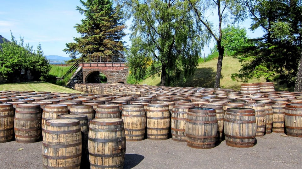 Holzfässer zur Whiskyreifung lagern im Freien auf einem Brennereigelände neben Brücke und Bäumen