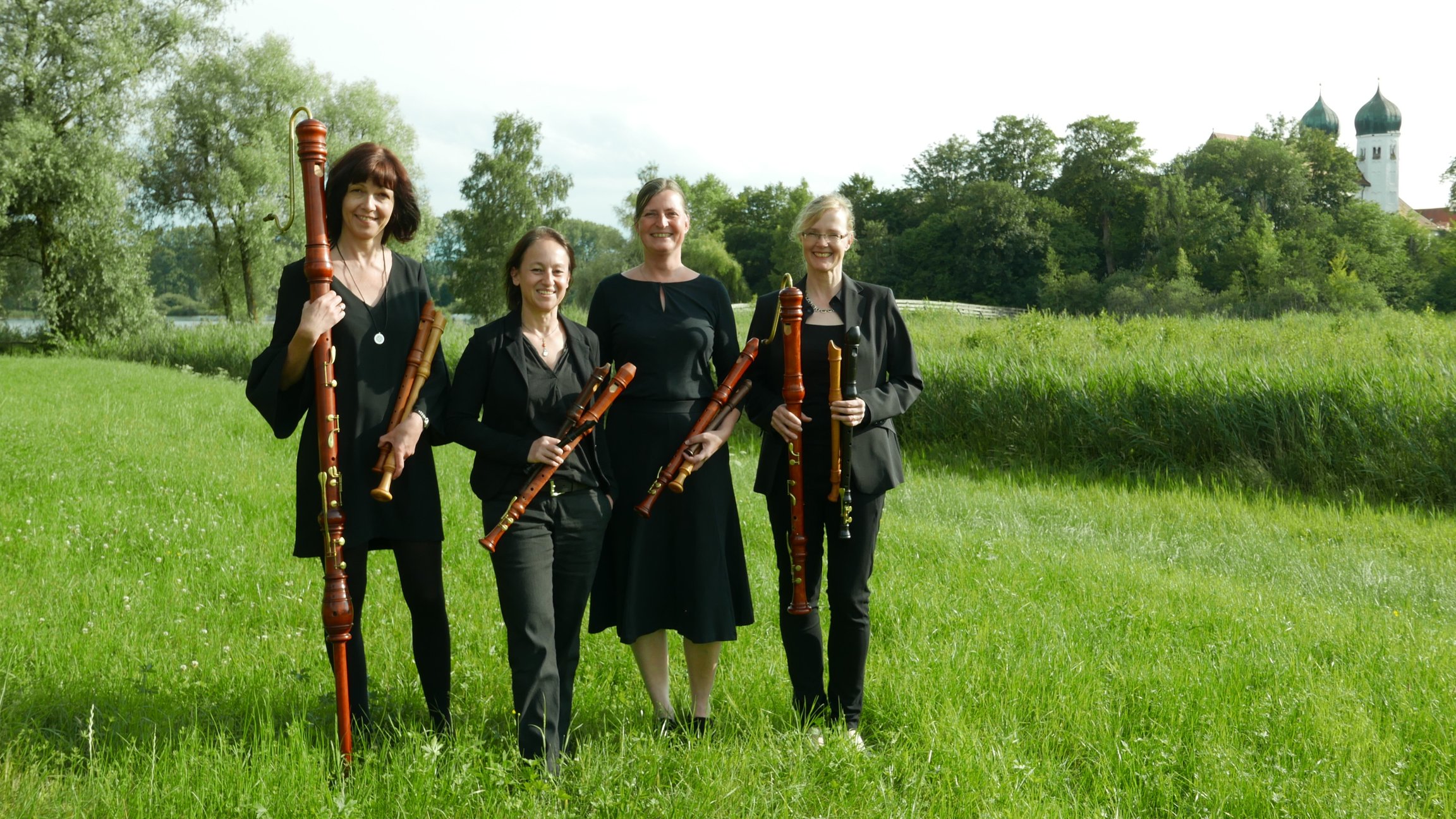 Blockflötenquartett Concerto Dolce steht im Grünen mit vier Musikerinnen in schwarzer Kleidung, die verschiedene Blockflöten in den Händen halten, im Hintergrund Wiese, Bäume und ein Gebäude mit Türmen.