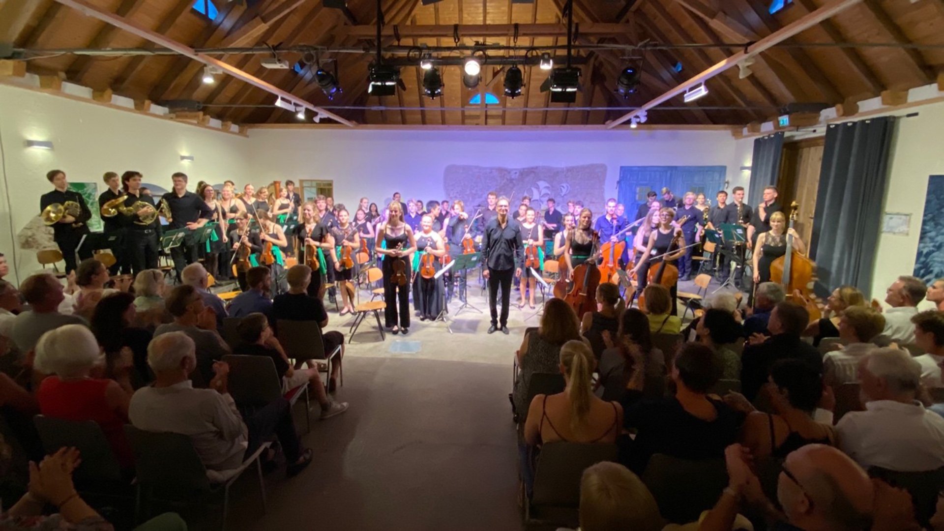Orchester mit Dirigenten steht nach einem Konzert vor sitzendem Publikum in einem Saal mit Holzbalkendecke.