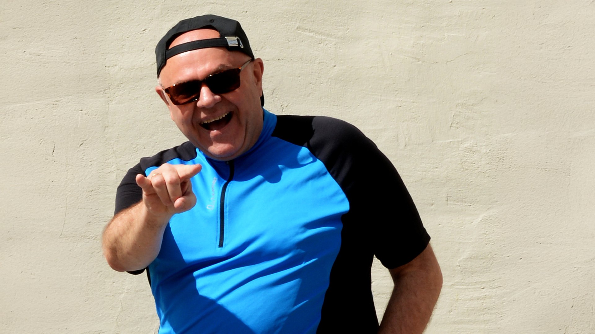 Foto des Künstlers Toni Lauerer mit einem blau-schwarzen T-Shirt, mit Mütze und Sonnenbrille vor einem hellen Hintergrund