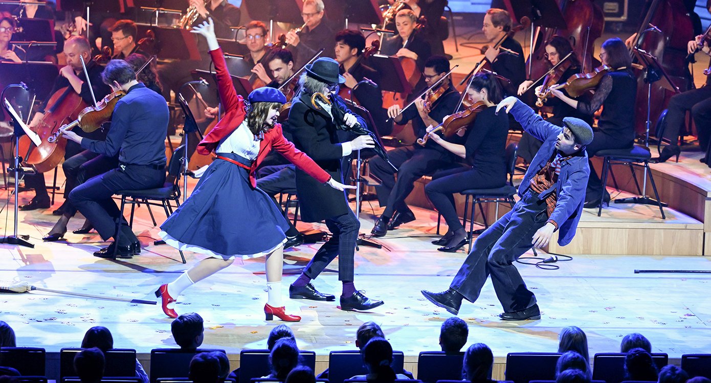 Ein Orchester spielt auf einer Bühne, während drei Schauspieler:innen in bunten Kostümen ein Theaterspiel für Kinder aufführen