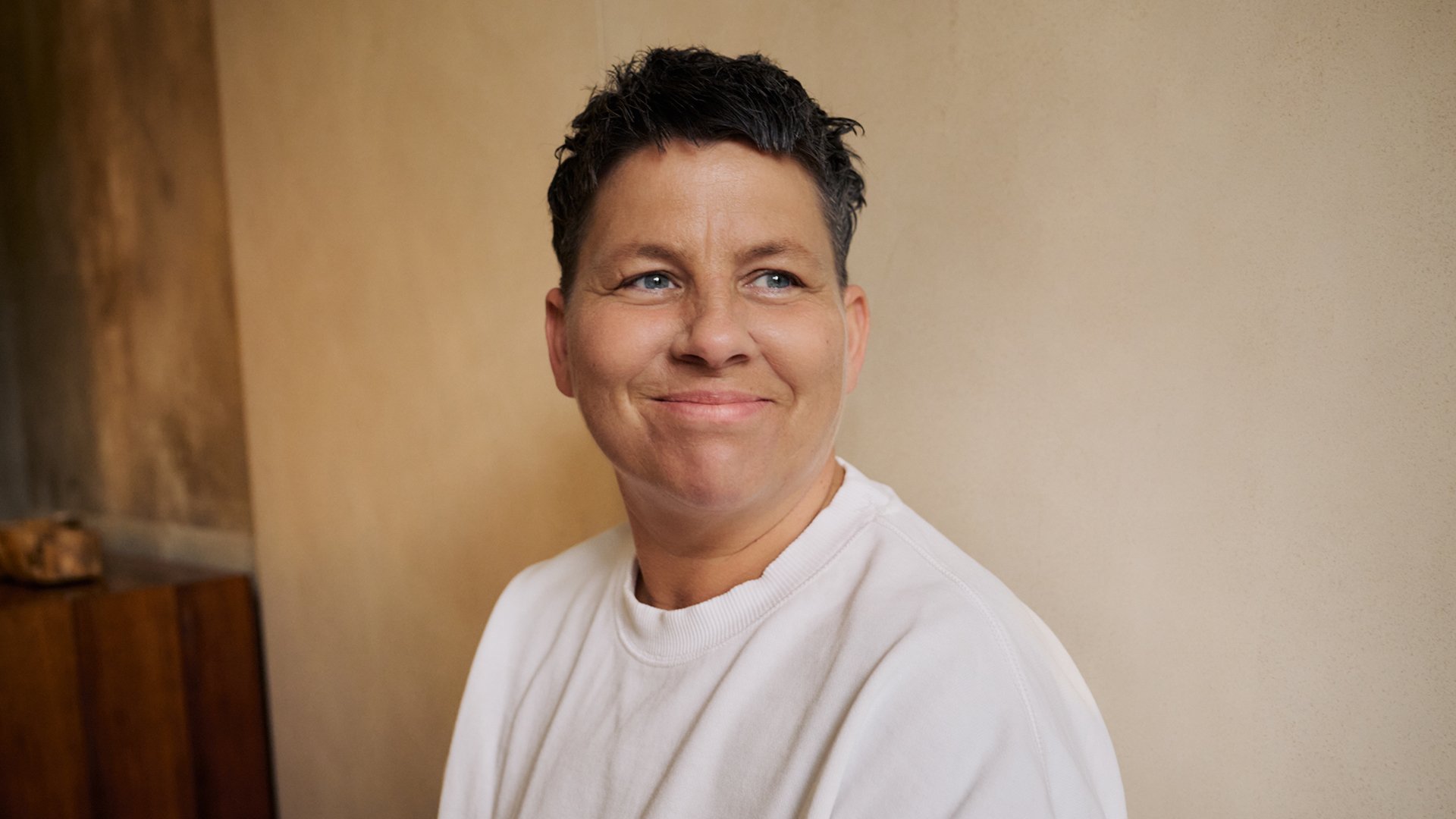 Kerstin Ott in weißem T-Shirt lächelnd, vor einer hellen Wand, auf der linken Seite eine Holzvertäfelung