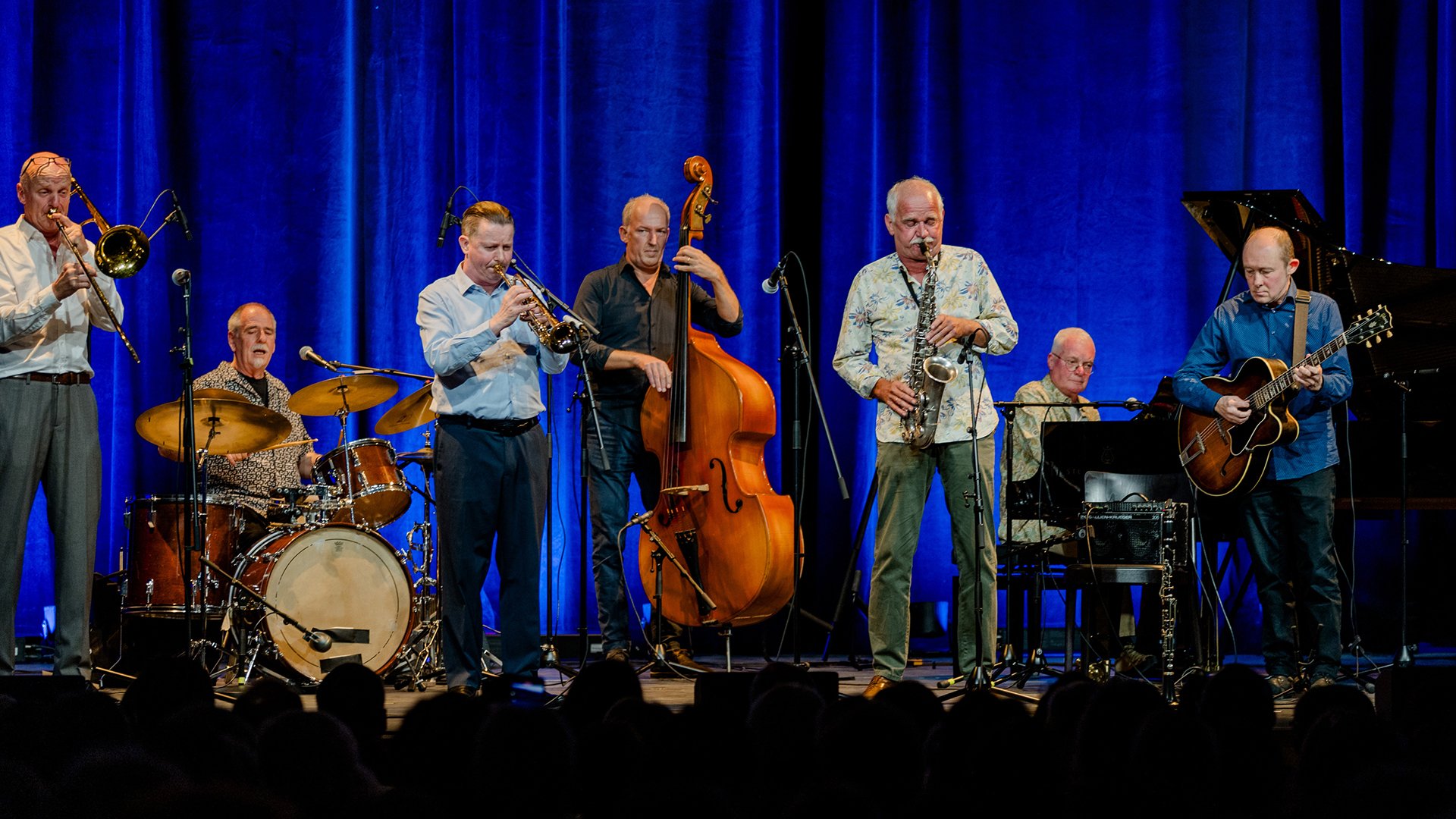 Bühnenfoto einer Jazzband mit sechs Musikern auf einer Bühne vor blauem Vorhang, darunter Posaune, Trompete, Saxofon, Kontrabass, Schlagzeug, Klavier und Gitarre, während des gemeinsamen Auftritts vor Publikum.