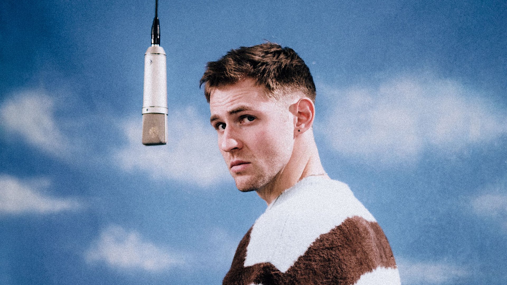 Dominik Hartz steht in braun-weiß getreiftem Pulloer vor blauem Himmel neben einem hängenden Studiomikrofon und blickt zur Kamera