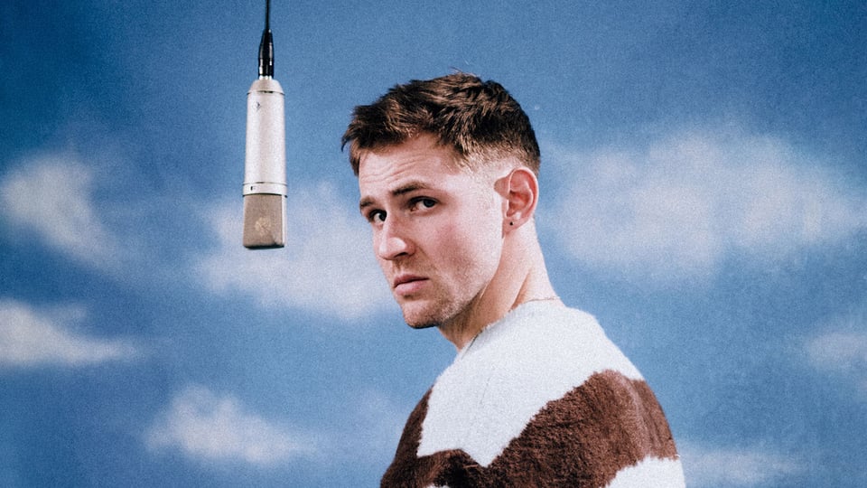 Dominik Hartz steht in braun-weiß getreiftem Pulloer vor blauem Himmel neben einem hängenden Studiomikrofon und blickt zur Kamera