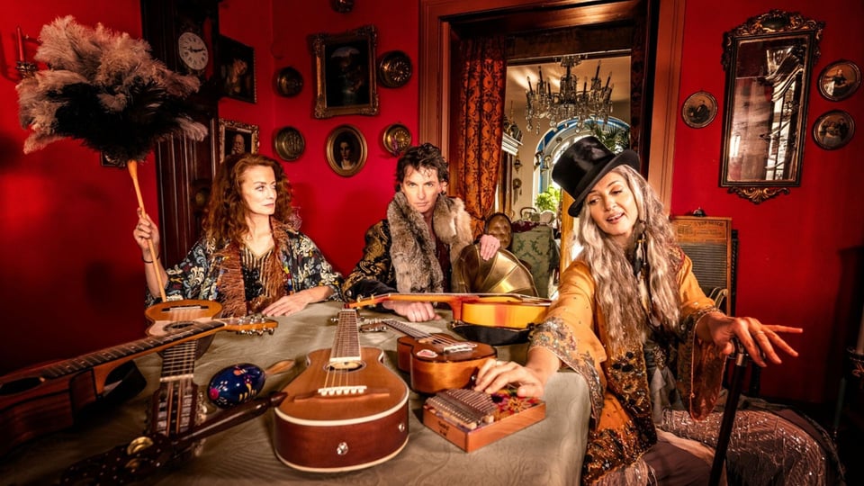 Drei Personen sitzen in einem aufwendig dekorierten Raum mit roten Wänden und Vintage‑Einrichtung, vor ihnen liegen verschiedene Musikinstrumente wie Gitarren und kleine Percussion‑Instrumente