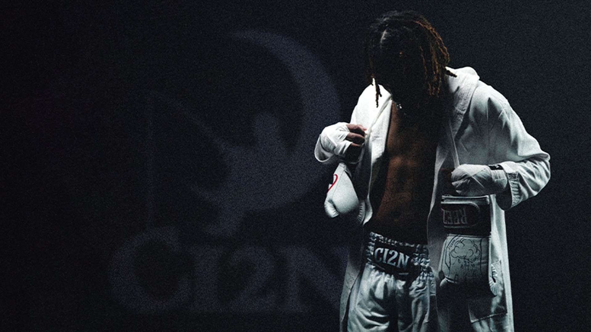 Reezy steht in einem dunklen Raum und trägt weiße Boxhandschuhe, weiße Boxershorts und einen offenen weißen Bademantel. 