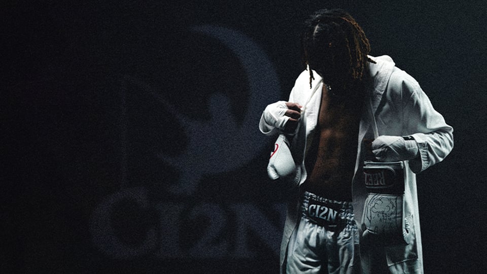 Reezy steht in einem dunklen Raum und trägt weiße Boxhandschuhe, weiße Boxershorts und einen offenen weißen Bademantel. 