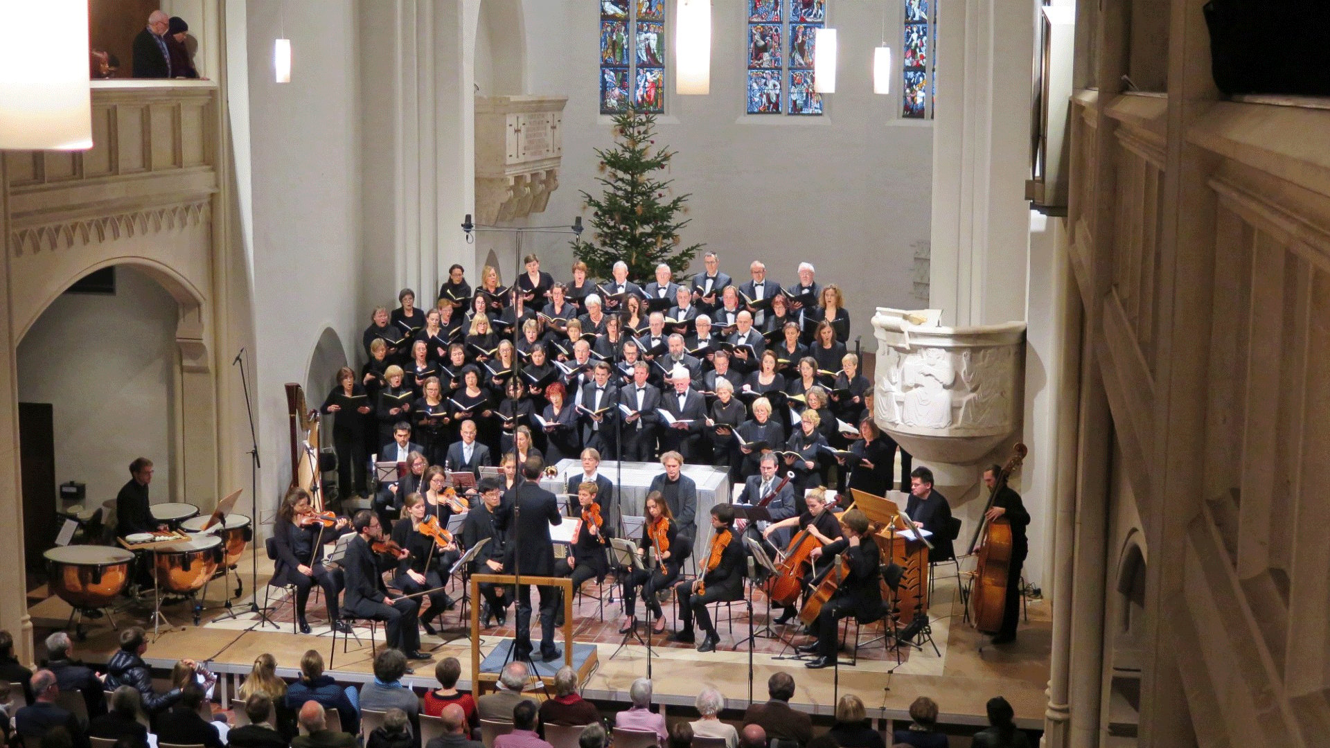 Konzertaufführung eines Chors mit Orchester in einer Kirche. Vor einem großen Chor auf mehreren Podesten dirigiert eine Person das Orchester mit Streichinstrumenten, Pauken und Kontrabass. Im Hintergrund sind ein Weihnachtsbaum sowie farbige Kirchenfenster zu sehen.