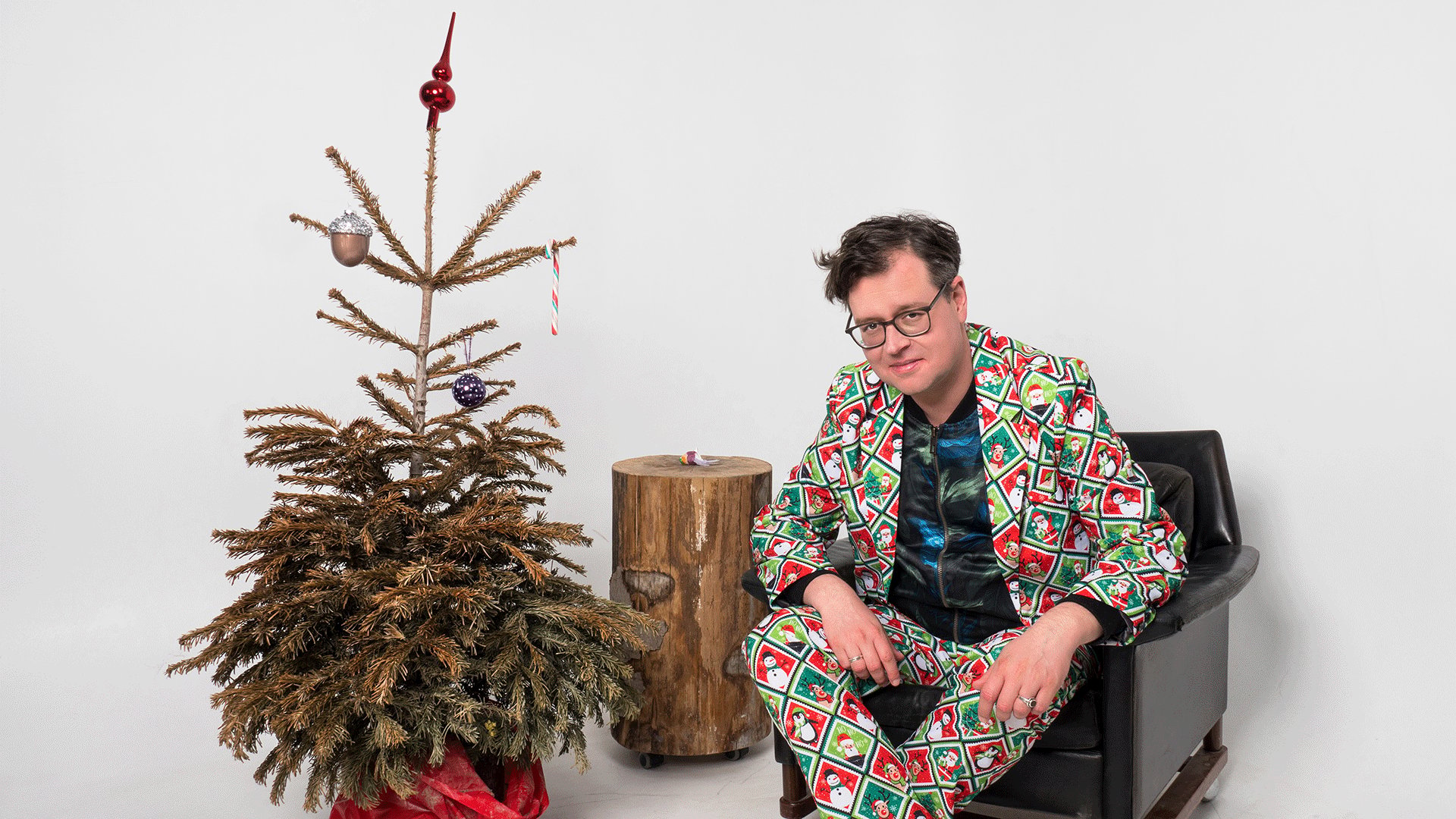 Der Comedian sitzt mit auffälligem Weihnachtsanzug in einem schwarzen Sessel neben einem vertrockneten Weihnachtsbaum mit spärlichem Baumschmuck vor weißem Hintergrund.