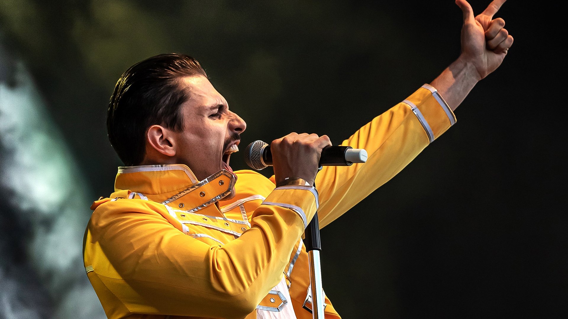 Der Darsteller von Freddy Mercury mit Mikrofon und gelbem Bühnenoutfit mit einer Handbewegung