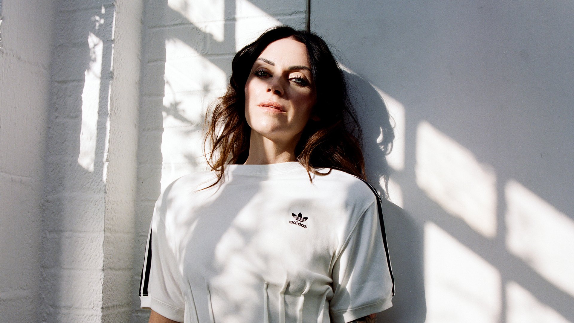 Porträtfoto von Amy Macdonald in weißem T-Shirt vor heller Wand, mit langen dunklen Haaren und Sonnenlicht, das durch Fensterrahmen Schatten auf Gesicht und Kleidung wirft