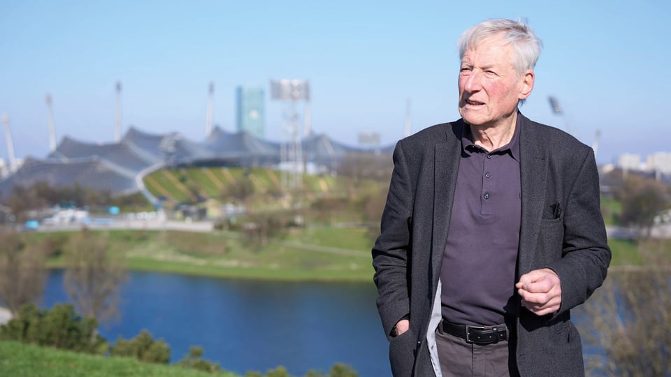 Person steht im Freien vor einer StadtlandArchitekt Prof. Fritz Auer steht im Freien vor dem Olympiagelände in München mit See und Stadion im Hintergrund bei sonnigem Wetterschaft mit See und Olympiastadion im Hintergrund, bei sonnigem Wetter