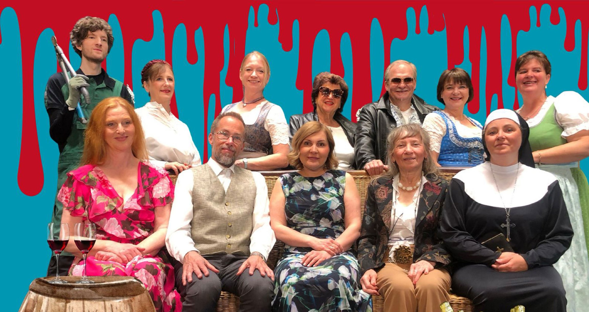 Ein Gruppenfoto der Schauspieler – einige sitzen, andere stehen dahinter. Daneben steht ein Tisch mit zwei Weingläsern.