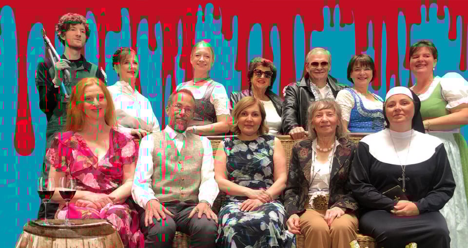 Ein Gruppenfoto der Schauspieler – einige sitzen, andere stehen dahinter. Daneben steht ein Tisch mit zwei Weingläsern.