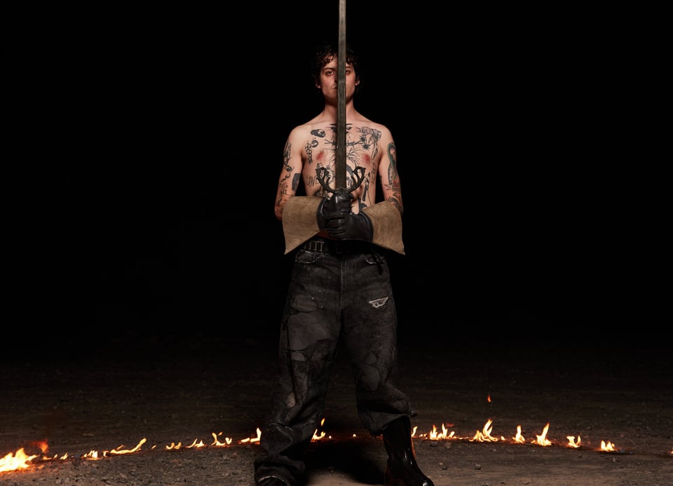 Foto des Künstlers mit einem Schwert in den Händen und einem Kreis aus Feuer mit kleinen Flammen auf dem Boden, vor schwarzem Hintergrund.