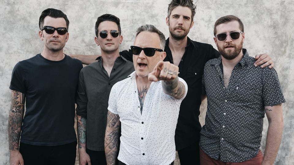 Dave Hause in weißem Hemd zeigt mit dem Zeigefindger in die Kamera und steht inmitten vier weiterer Personen