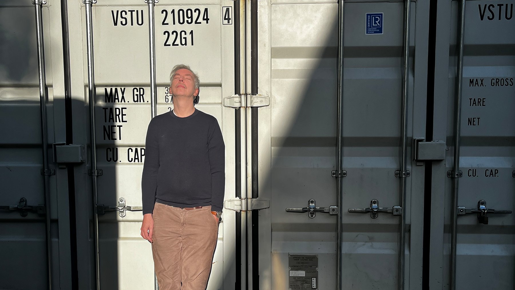 Olli Schulz im grauen Pullover und heller Hose steht mit geschlossenen Augen im Sonnenlicht vor grauen Containern, ein teil liegt im Schatten
