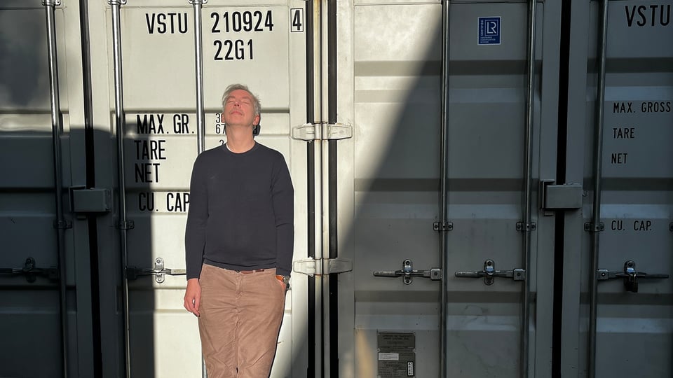 Olli Schulz im grauen Pullover und heller Hose steht mit geschlossenen Augen im Sonnenlicht vor grauen Containern, ein teil liegt im Schatten